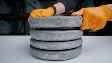Hands in orange gloves stacking gray metal alloy slabs on a white surface, forming a tower.