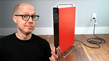 Man with glasses sits at left of a bright red, tall appliance plugged into a wall outlet on a wooden floor, gesturing with hi