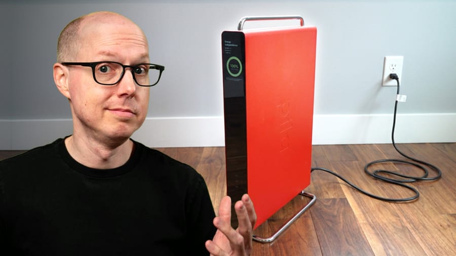 Man with glasses sits at left of a bright red, tall appliance plugged into a wall outlet on a wooden floor, gesturing with hi